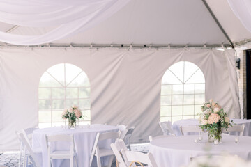 A tented backyard wedding reception with white tables and simple flower arrangement in the center of each table. 