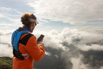 A man in an orange jacket is looking at his cell phone while standing on a mountain. The sky is...