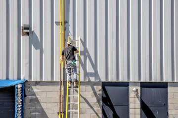 Man standing on a ladder painting natural gas lines yellow on the side of a building with grey...
