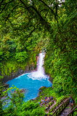 Fototapeta premium A vibrant waterfall cascades into a turquoise pool surrounded by lush green rainforest. A group of tourists descends a rustic staircase to view the natural beauty up close. High quality photo. Rio