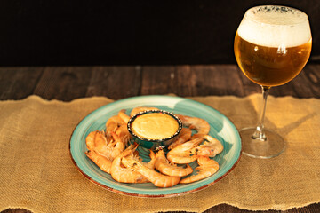 Boiled prawns with sauce and a glass of light beer served on a wooden table with canvas cloth.