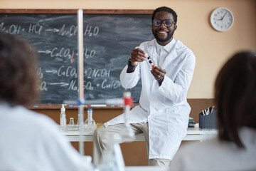 Over-the-shoulder shot of cheerful young African American teacher of Chemistry wearing white lab...
