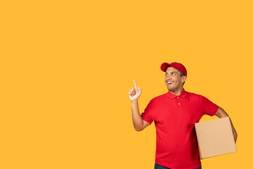 A cheerful guy delivery person dressed in a red uniform points upward with a confident smile while holding a package. The background is a vibrant yellow, copy space