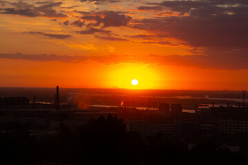 Beautiful panoramic cityscape of industrial district during romantic sunrise. Orange sky and clouds. Soft focus. Silhouettes of manufacturing buildings with pipes by river. City landscape theme.
