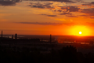 Beautiful panoramic cityscape of industrial district during romantic sunrise. Orange sky and clouds. Soft focus. Silhouettes of manufacturing buildings with pipes by river. City landscape theme.
