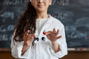 Medium closeup of schoolgirl wearing lab coat standing against blackboard holding chemical structure model in hands