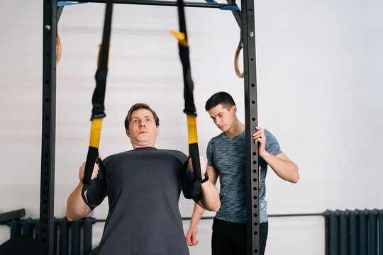 Determined sporty beginner sportsman practicing suspension training using sling trainer with male fitness instructor, doing pull-up and bodyweight exercise in health club, physical sport activity