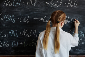 Rear view of unrecognizable schoolgirl wearing white lab coat writing formulae on blackboard in...