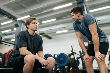 Low-angle view of of muscular personal instructor explaining technique of exercise to young beginner sportsman sitting on bench with barbell during fitness training. Concept of healthy life.