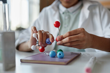 Closeup of hands of unrecognizable Black schoolboy wearing white lab coat sitting at desk assembling molecular structure model