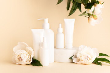 Set of skin care cosmetic products in white plastic unbranded containers on light beige background decorated with aromatic white peony flowers.