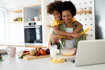 Playful young girl embracing her loving mother and holding spaghetti straps in both hands while making pasta for lunch, whipping ingredients in a bowl and using laptop for online cooking recipes.