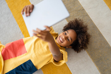 Smiling African American girl using digital tablet while lying on the floor at home.