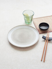Table set for oriental food. Ceramic plate and green glass on the table, wooden chopsticks and small black bowl.