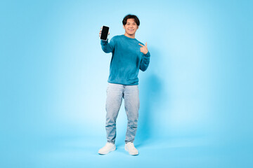 Asian young guy is standing in a brightly lit studio, smiling and pointing to a smartphone he is holding in his left hand. The vibrant blue background