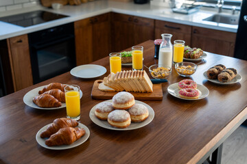 Food served on the dining table. Festive brunch set, meal variety with donuts, croissants, sandwiches, milk and orange juice. Brunch at home. Breakfast in the kitchen. No people on the photo.