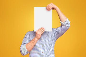 A man holding a blank white piece of paper over his face in the air against a plain yellow studio...