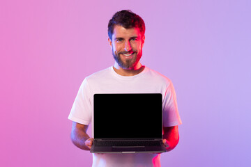 A young man in a white t-shirt smiles while holding an open laptop with black screen in a studio....