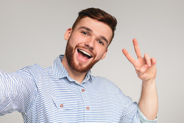 Young millennial man wearing casual shirt showing V - sign and making selfie on grey studio...
