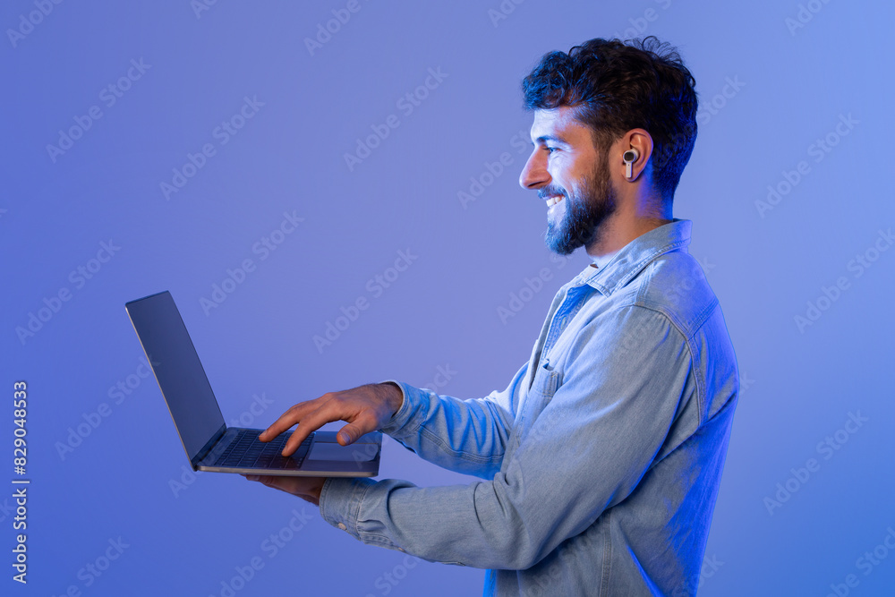 Poster A man with a beard is sitting with a laptop open in front of him. He appears focused as he types on the keyboard, possibly working or studying.