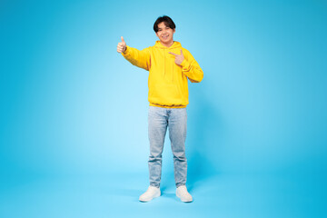 Asian guy wearing a bright yellow hoodie is enthusiastically giving a thumbs up gesture. He appears...