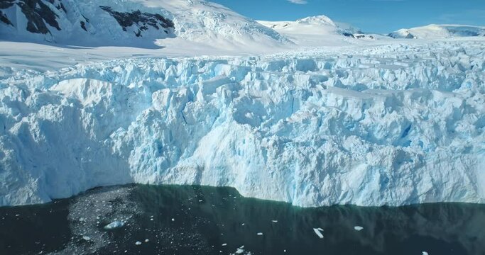 Zoom out panorama giant glacier ice wall iceberg towering on Antarctic coastline polar ocean. Sunny day blue sky, snow covered mountains. Ecology, global warming and climate change. Aerial drone shot