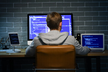 Hacker using laptop at table in dark room, back view