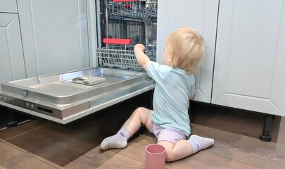 Baby toddler is standing by an open dishwasher, child puts glass in dishwasher.Child safety issues...