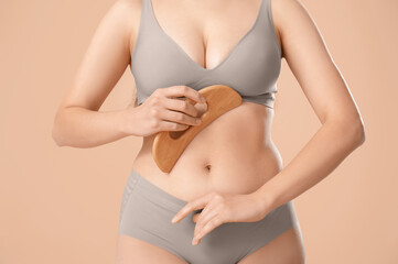 Young woman in underwear massaging her belly with wooden scraper on beige background, closeup