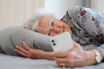 Senior woman lying in bed, looking at smartphone and smiling. Older woman using technology, digital...