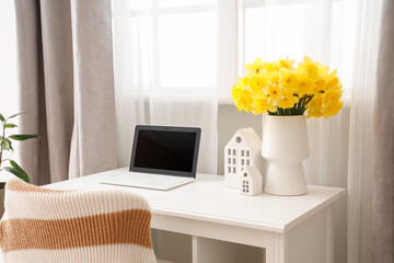 Workplace with laptop and vase of narcissus flowers in stylish room