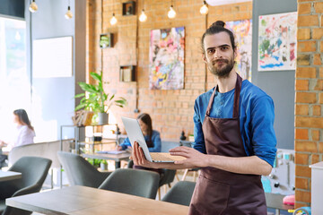 Fototapeta premium Man service worker owner in apron using laptop in restaurant cafeteria interior