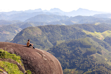 Man sitting on the edge of a cliff, gazing at mountains in the background. Ideal for outdoor enthusiasts seeking stress relief or pursuit of personal growth  through travel and adventurous experiences