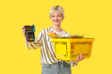 Young woman with full shopping basket and payment terminal on yellow background