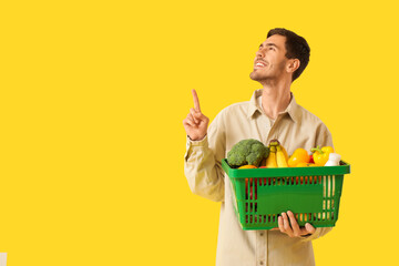 Young man with full shopping basket pointing at something on yellow background