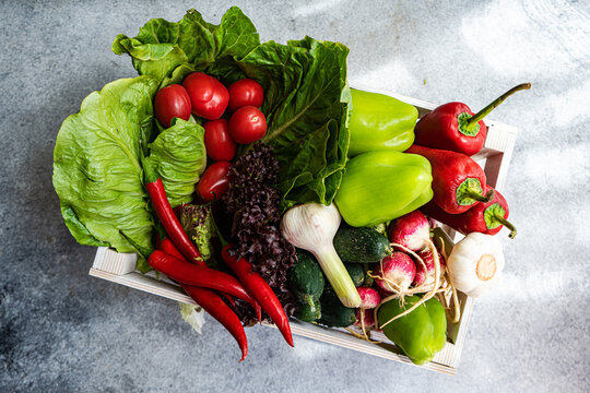 Overhead view of a wooden crate filled with fresh lettuce, bell peppers, chillis, tomatoes, radishes, cucumber and garlic