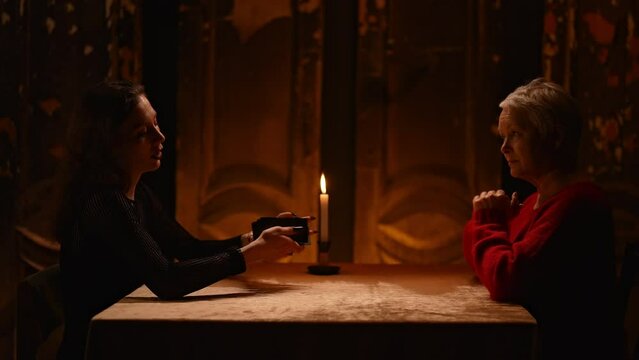 Side wide shot of mysterious fortune teller giving spiritual session with tarot card for adult female client sitting at table in dark esoteric room by light of burning candle. Shooting in slow motion.