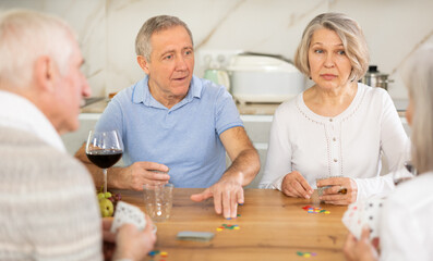 Two senior married couples are having fun in cozy home atmosphere, playing card game poker. Player makes lead move and believes in luck, emotionally waiting for victory, calculate winning combination