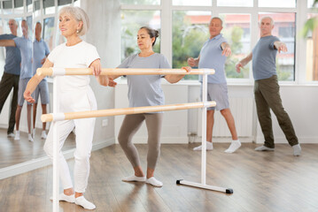 Elderly active women perform various ballet positions at the barre, men in the background perform...