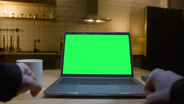 Point of view shot: Man uses laptop computer with green mock up screen being happy, celebrate winning in the modern kitchen background at night.