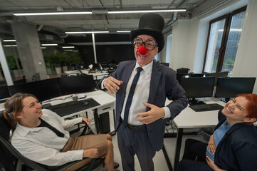 An elderly Caucasian man in a clown costume amuses two Caucasian women in the office. 