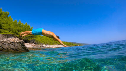 LOW ANGLE VIEW: Young man in shorts jumps head first from rocky shore and dives into crystal clear...