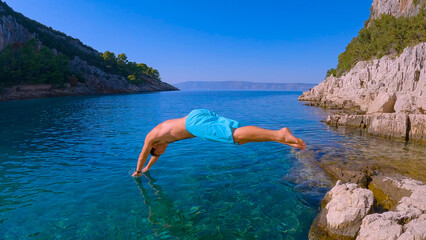 Active young tourist launches himself head first from a raised rock into incredibly clear deep blue...