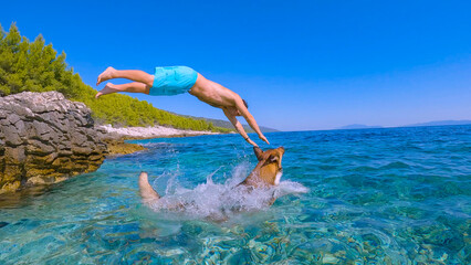 LOW ANGLE VIEW: Young guy and his loyal pet enjoy jumping into refreshing sea. Splashing dip from...