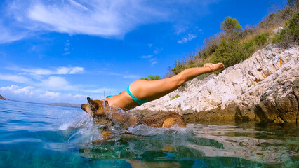 LOW ANGLE VIEW: Woman and her doggo jump off a rugged shore into crystal blue sea. Refreshing dive...