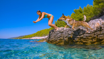 LOW ANGLE VIEW: Woman and her mixed breed dog jump off a rock into the blue sea to refresh...