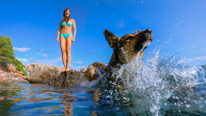 LOW ANGLE VIEW: Lady in bikini stands on sea rock when her dog jumps into water. Upon landing in...