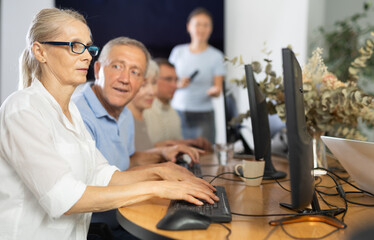 Old-aged female student learning computer in group and teacher standing on the background