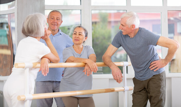 Bouyant senior male and female participants of ballet classes chatting joyfully standing around training barre