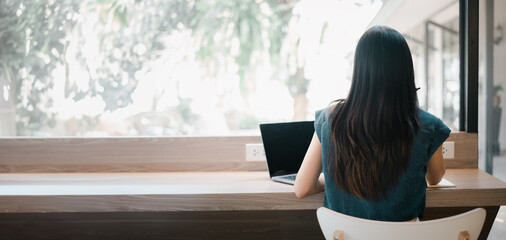 A woman sits at a desk with a laptop in front of her. She is wearing a blue shirt and has long...